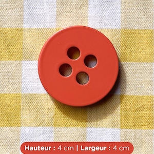 Peut inclure: Un gros bouton rond orange avec quatre trous, d'environ 4 cm de diamètre. Le bouton est posé sur un fond de tissu à carreaux jaunes et blancs. Le texte sur l'image indique : Hauteur : 4 cm | Largeur : 4 cm.