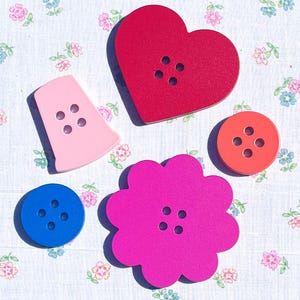 Peut inclure: Un ensemble de cinq boutons de formes et de couleurs variées, comprenant un grand cœur rouge, une fleur rose vif, un petit bouton rond orange, un petit bouton rond bleu et un bouton rose clair en forme de dé à coudre, chacun avec quatre trous.
