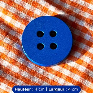 Peut inclure: Un gros bouton rond bleu vif avec quatre trous. Le bouton mesure 4 cm de hauteur et 4 cm de largeur. Le bouton est posé sur un tissu à carreaux orange et blanc.