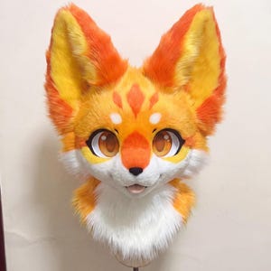 以下が含まれることがあります： 大きく表現力豊かな目を備えた、鮮やかなオレンジと黄色のキツネのファースーツヘッド。毛皮の頭には白いマズルと胸があり、顔のマーキングが施されています。耳は直立しており、全体的なデザインは漫画的でフレンドリーです。