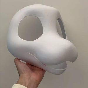 Może przedstawiać: Biała, drukowana w 3D baza głowy fursuit z dużymi otworami na oczy i pyskiem. Głowa trzymana jest przez dłoń, ukazując gładką, niepomalowaną powierzchnię. Projekt sugeruje stylizowane zwierzę lub postać.