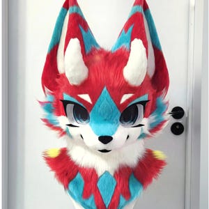 Może przedstawiać: Kolorowa głowa fursuit z lisim designem. Głowa jest głównie czerwona z niebieskimi i białymi akcentami, w tym uszami, oczami i oznaczeniami na twarzy. Fursuit ma duże, wyraziste oczy i biały pysk.