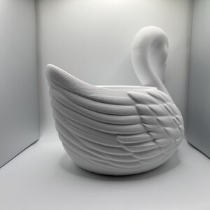 May include: A matte white swan-shaped planter or decorative bowl featuring detailed molded feather textures along the body and a curved neck. The object has a smooth, uniform finish and a hollow center, showcasing a sculptural design.