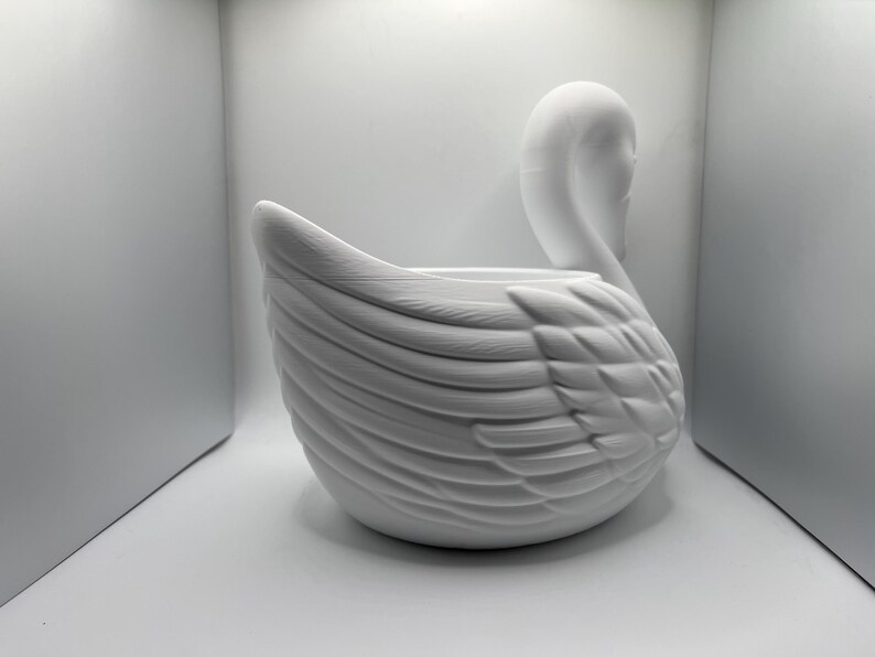 May include: A matte white swan-shaped planter or decorative bowl featuring detailed molded feather textures along the body and a curved neck. The object has a smooth, uniform finish and a hollow center, showcasing a sculptural design.