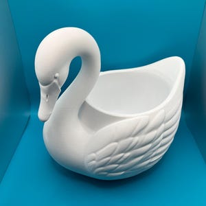 May include: A matte white ceramic swan-shaped planter or decorative bowl. The swan features a curved neck, a detailed beak, and molded feather textures along the body, with a hollow, open center for holding items or small plants.