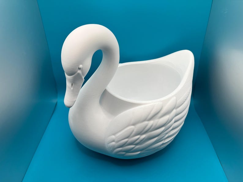 May include: A matte white ceramic swan-shaped planter or decorative bowl. The swan features a curved neck, a detailed beak, and molded feather textures along the body, with a hollow, open center for holding items or small plants.
