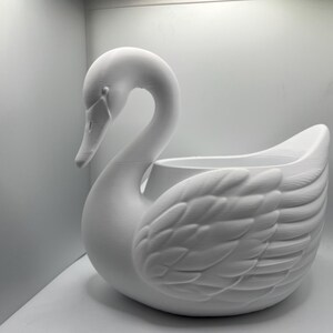 May include: A matte white swan-shaped planter featuring a long, curved neck and detailed molded feather textures on the wings. The body of the swan forms a hollow basin, providing an open space for holding items or plants within its structure.