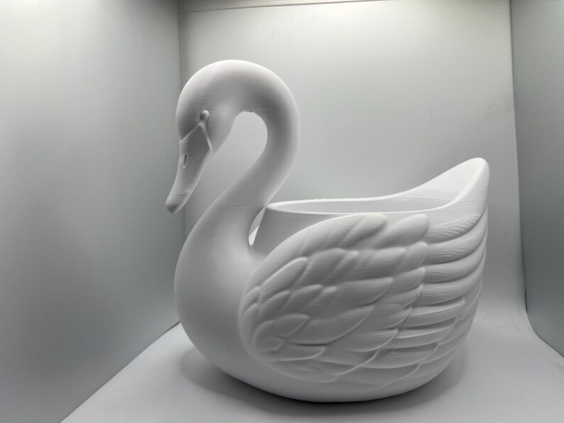 May include: A matte white swan-shaped planter featuring a long, curved neck and detailed molded feather textures on the wings. The body of the swan forms a hollow basin, providing an open space for holding items or plants within its structure.