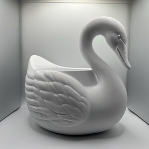 May include: A matte white swan-shaped planter featuring a curved neck and detailed wing feathers. The hollow body provides an open space for plants or decorative items, showcasing a smooth, uniform finish across the entire swan structure.
