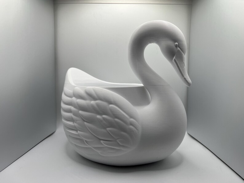 May include: A matte white swan-shaped planter featuring a curved neck and detailed wing feathers. The hollow body provides an open space for plants or decorative items, showcasing a smooth, uniform finish across the entire swan structure.
