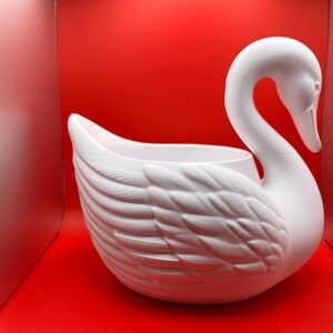 May include: A matte white swan-shaped planter or decorative bowl featuring a long, curved neck and detailed, layered feather textures on the body. The swan has a hollow center, creating an open basin for holding items, with a smooth, clean finish.