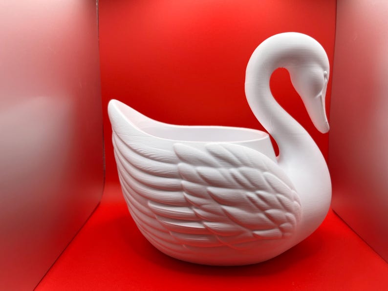 May include: A matte white swan-shaped planter or decorative bowl featuring a long, curved neck and detailed, layered feather textures on the body. The swan has a hollow center, creating an open basin for holding items, with a smooth, clean finish.