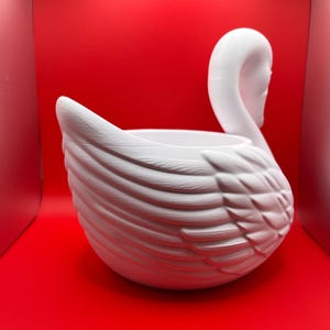 May include: A white swan-shaped planter featuring a textured feather design on the exterior. The swan has a long, curved neck and a hollow center, creating a basin for holding items. The surface has a matte finish with visible molded details.