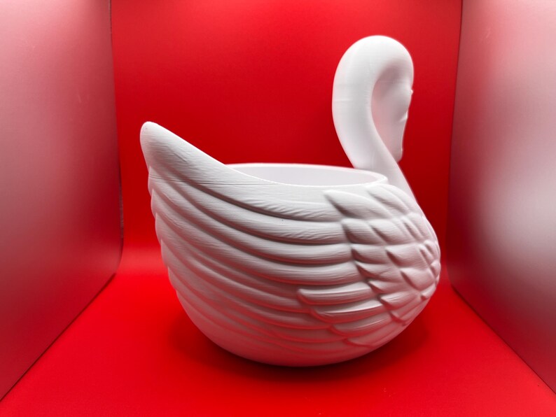 May include: A white swan-shaped planter featuring a textured feather design on the exterior. The swan has a long, curved neck and a hollow center, creating a basin for holding items. The surface has a matte finish with visible molded details.