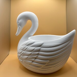 May include: A matte white swan-shaped planter featuring a long, curved neck and detailed molded feathers on the body. The center of the swan has a hollow, circular opening designed for holding items, with a smooth interior and a textured exterior.