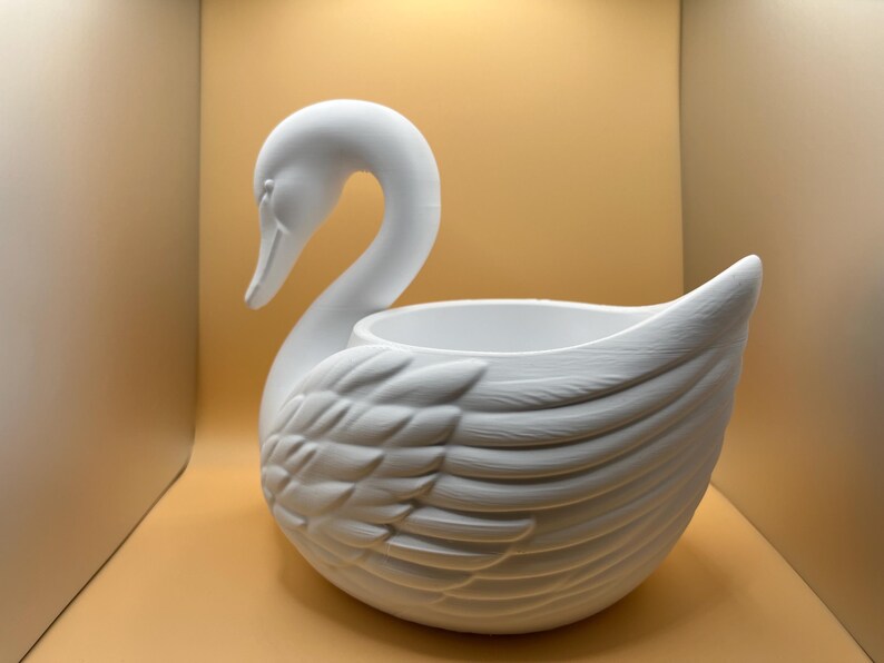 May include: A matte white swan-shaped planter featuring a long, curved neck and detailed molded feathers on the body. The center of the swan has a hollow, circular opening designed for holding items, with a smooth interior and a textured exterior.