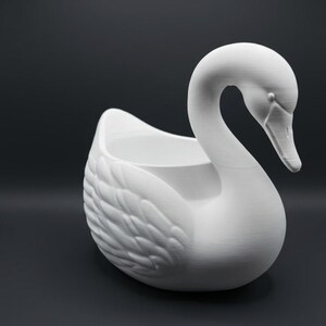 May include: A matte white swan-shaped planter or decorative container featuring a curved neck and detailed feather textures on the wings. The hollow center is open at the top, and the swan is depicted in a resting position with its head tilted downward.