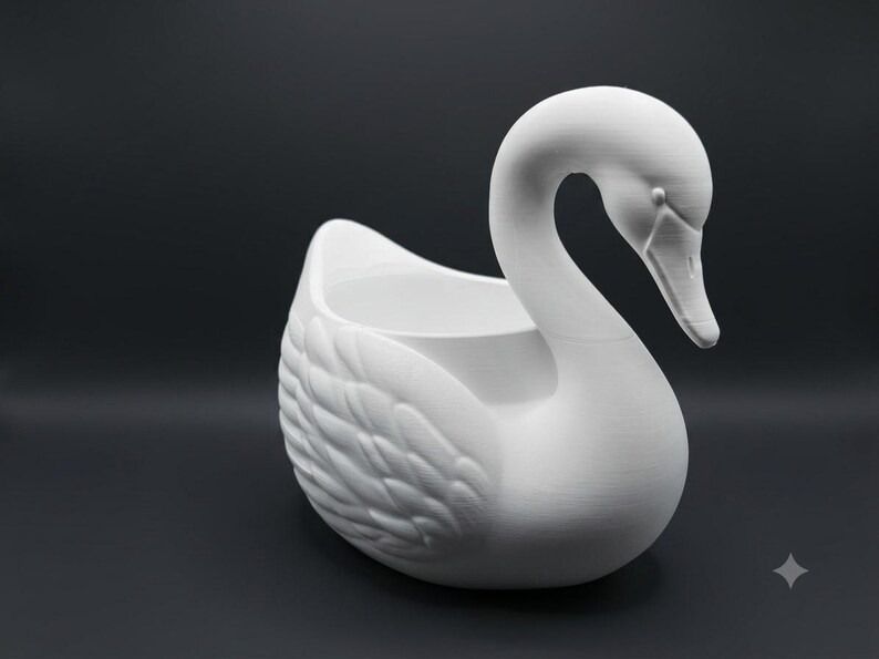 May include: A matte white swan-shaped planter or decorative container featuring a curved neck and detailed feather textures on the wings. The hollow center is open at the top, and the swan is depicted in a resting position with its head tilted downward.