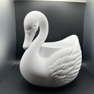 May include: A matte white swan-shaped planter featuring a long, curved neck and detailed molded feather textures on the wings. The hollow center provides an open basin for contents, and the entire structure is finished in a solid, uniform white.