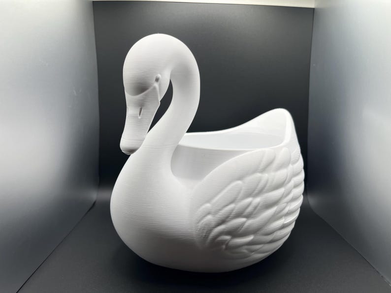 May include: A matte white swan-shaped planter featuring a long, curved neck and detailed molded feather textures on the wings. The hollow center provides an open basin for contents, and the entire structure is finished in a solid, uniform white.
