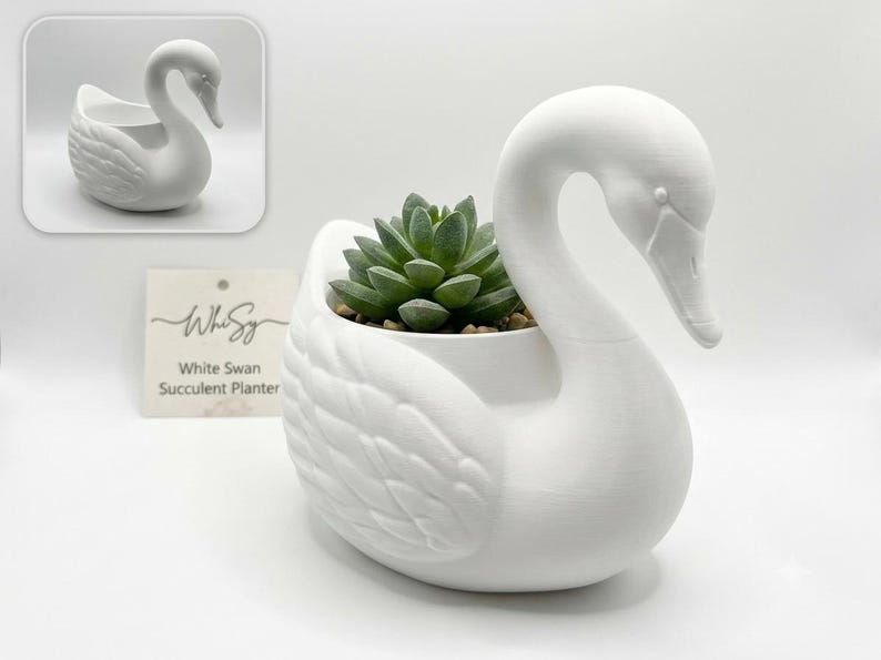 May include: A matte white swan-shaped planter containing a green succulent plant. The swan features detailed wing textures and a long, curved neck. A small square card is visible in the background with the text WhiSy White Swan Succulent Planter.