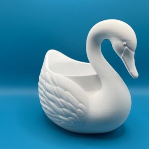 May include: A matte white swan-shaped planter featuring a hollow center for holding items. The swan has a long, curved neck, a defined beak, and textured wing details on its sides. The object is presented against a solid blue background.