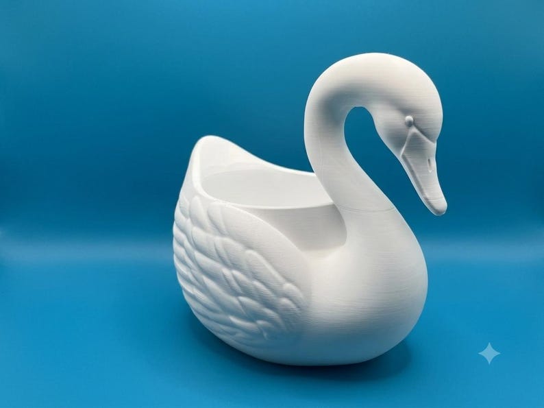 May include: A matte white swan-shaped planter featuring a hollow center for holding items. The swan has a long, curved neck, a defined beak, and textured wing details on its sides. The object is presented against a solid blue background.