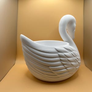 May include: A matte white ceramic planter shaped like a swan. The vessel features a hollow center for planting and detailed, molded feather textures along the exterior body, with the swan's neck and head curved gracefully upward from the side.