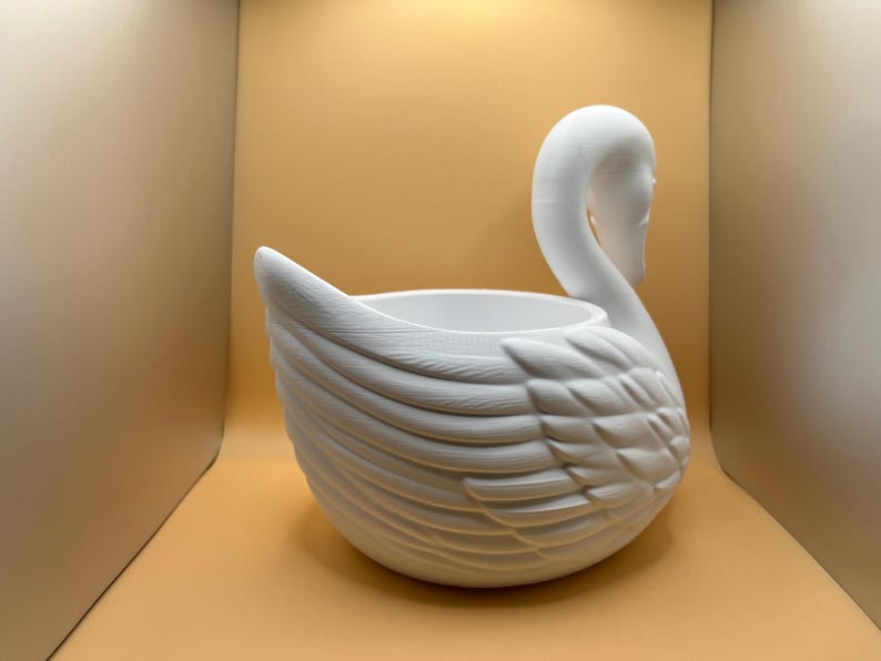 May include: A matte white ceramic planter shaped like a swan. The vessel features a hollow center for planting and detailed, molded feather textures along the exterior body, with the swan's neck and head curved gracefully upward from the side.