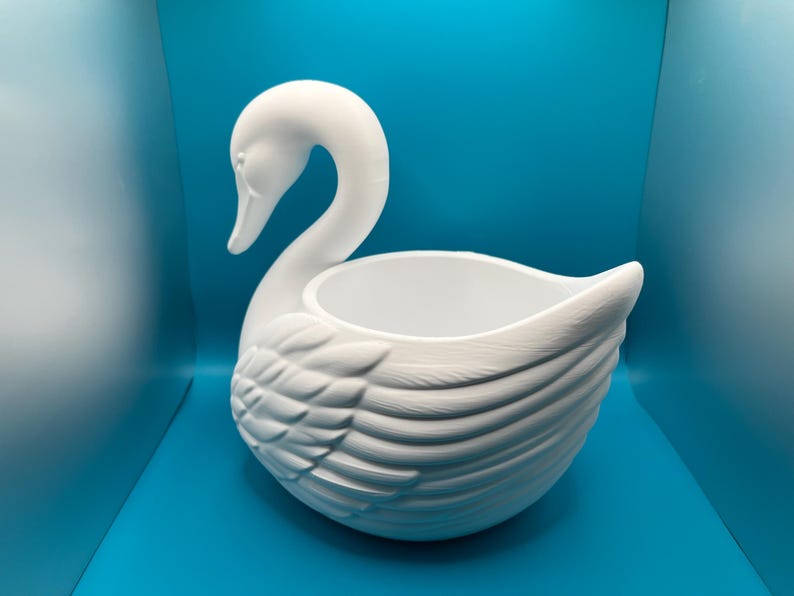 May include: A matte white swan-shaped planter or decorative bowl featuring a long, curved neck and detailed molded feather textures on the wings. The interior is smooth and hollow, providing an open basin for holding small items or plants.