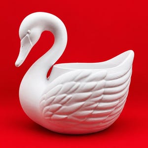 May include: A matte white swan-shaped planter or decorative bowl featuring a long, curved neck and detailed, textured wing feathers. The object has a hollow center and is positioned against a solid, vibrant red background.