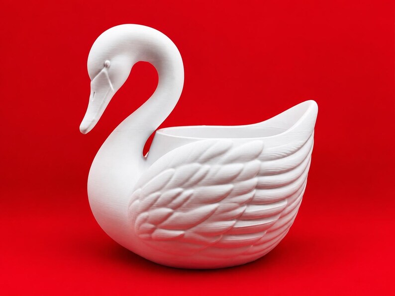 May include: A matte white swan-shaped planter or decorative bowl featuring a long, curved neck and detailed, textured wing feathers. The object has a hollow center and is positioned against a solid, vibrant red background.