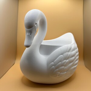 May include: A matte white swan-shaped planter featuring a smooth, curved neck and detailed molded wing feathers on the sides. The hollow center provides an open space for contents, while the swan head is positioned facing downward toward the front.