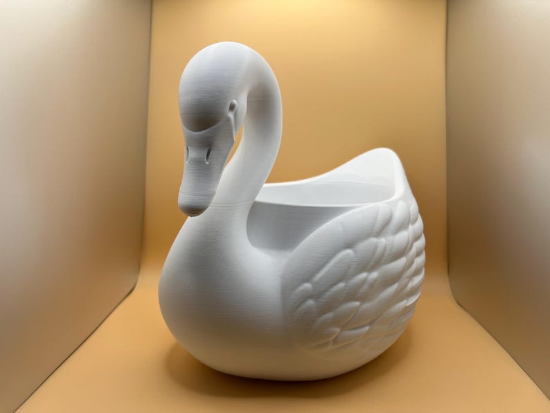 May include: A matte white swan-shaped planter featuring a smooth, curved neck and detailed molded wing feathers on the sides. The hollow center provides an open space for contents, while the swan head is positioned facing downward toward the front.