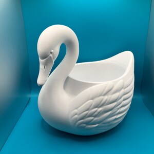 May include: A matte white ceramic swan-shaped planter or decorative bowl. The swan features a long, curved neck, a detailed beak, and molded feather textures along the body. The center of the swan is hollow, creating an open basin for storage.