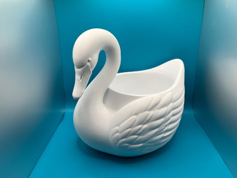 May include: A matte white ceramic swan-shaped planter or decorative bowl. The swan features a long, curved neck, a detailed beak, and molded feather textures along the body. The center of the swan is hollow, creating an open basin for storage.