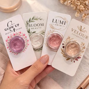 Puede incluir: Cuatro tarros de cosméticos con cremas de diferentes colores se muestran en tarjetas blancas. Las tarjetas tienen el siguiente texto: "Glow Up Studio Beauty", "Bloom Beauty", "Lumi Skin" y "Velour Cosmetics". Los tarros son sostenidos por una mano.