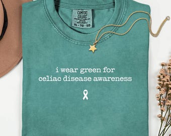 May include: Teal t-shirt with the text "i wear green for celiac disease awareness" and a white awareness ribbon. A gold star pendant necklace rests on the shirt. The shirt is a Comfort Colors XL-TG-EG.