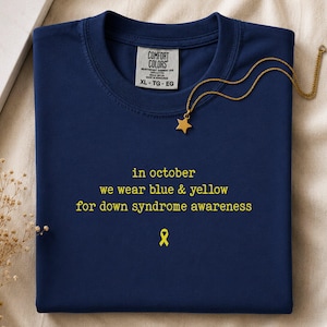 Op de afbeelding: Een marineblauw t-shirt met de gele tekst "in october we wear blue & yellow for down syndrome awareness" en een geel icoon van een bewustwordingslint. Een goudkleurige ketting met een kleine sterhanger rust op de kraag van het shirt.