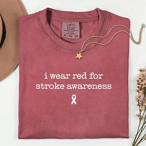 Op de afbeelding: Een rood t-shirt met de tekst "i wear red for stroke awareness" en een wit lintsymbool. Een gouden sterrenhangerketting ligt op het shirt. Het shirt is opgevouwen en heeft een label met "Comfort Colors".