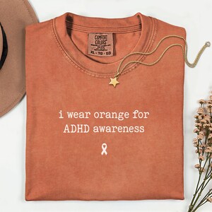 Pode incluir: Uma t-shirt cor de laranja queimado com uma etiqueta Comfort Colors no colarinho. A peça apresenta o texto branco "i wear orange for ADHD awareness" acima de um pequeno ícone de fita de consciencialização branca. Um colar de corrente dourada com uma estrela está colocado por cima.