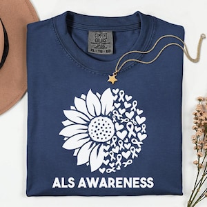 May include: A navy blue t-shirt featuring a white graphic of a sunflower on the left side and a collection of hearts and awareness ribbons on the right side. The text ALS AWARENESS is printed in white capital letters at the bottom of the shirt.