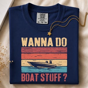 Peut inclure: T-shirt bleu marine avec l'inscription "WANNA DO BOAT STUFF?" dans un style rétro. Le graphisme comprend une illustration de bateau et des rayures horizontales colorées. Un collier doré avec un pendentif étoile est posé sur le t-shirt.