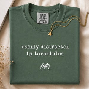 May include: A dark olive green crew neck t-shirt featuring the white text "easily distracted by tarantulas" and a small white spider graphic below. The shirt has a Comfort Colors brand label at the neck and is paired with a gold star pendant necklace.
