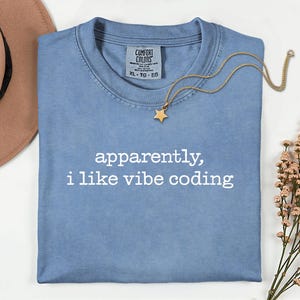 Op de afbeelding: Een lichtblauw T-shirt met de witte tekst "apparently, i like vibe coding". Een goudkleurige ketting met een sterhanger ligt op het shirt. Het shirt is op een wit oppervlak gevouwen.