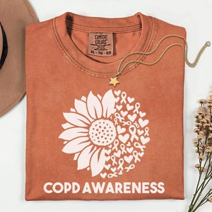 Może przedstawiać: T-shirt w kolorze rdzawym z białą grafiką przedstawiającą słonecznik po lewej stronie oraz zbiór serc i wstążek świadomości po prawej. Napis "COPD AWARENESS" jest nadrukowany białymi wielkimi literami w dolnej części koszulki.