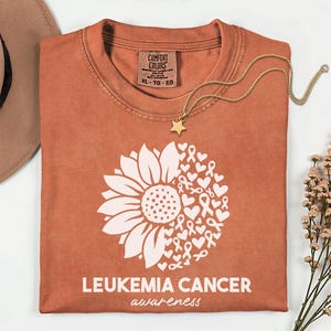 Könnte beinhalten: Ein rostfarbenes Kurzarm-T-Shirt mit einer weißen Grafik aus einer Sonnenblume, kombiniert mit Herzen und Bewusstseinsschleifen. Der Text LEUKEMIA CANCER awareness ist unter der Grafik aufgedruckt. Das Shirt hat ein Comfort Colors Etikett.