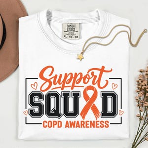 Può includere: Una t-shirt bianca con la scritta Support SQUAD e COPD AWARENESS in lettere arancioni e nere. Il design include un nastro di sensibilizzazione arancione e piccoli cuori. La maglietta ha un'etichetta Comfort Colors che indica la taglia XL.