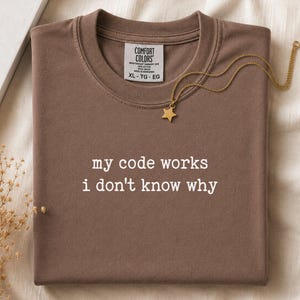 May include: A brown short-sleeve t-shirt with the white text "my code works i don't know why" printed on the chest. The shirt features a Comfort Colors brand label at the inner neckline and is accompanied by a gold-toned star pendant necklace.