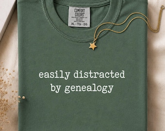 May include: A forest green crewneck t-shirt with the white text "easily distracted by genealogy" printed on the chest. The shirt features a Comfort Colors brand label at the inner neckline and is paired with a gold-toned star pendant necklace.